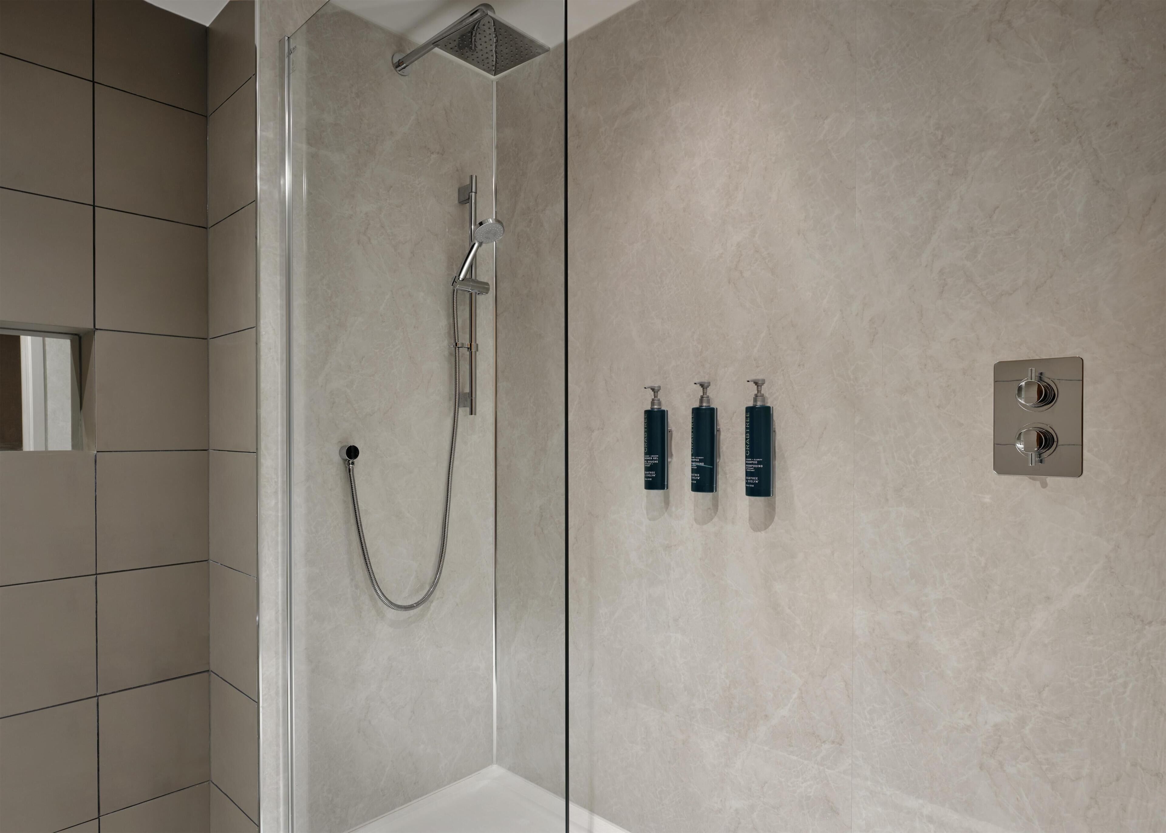 A modern shower with a glass enclosure, a handheld showerhead, and a wall-mounted control panel with three bottles of shampoo.