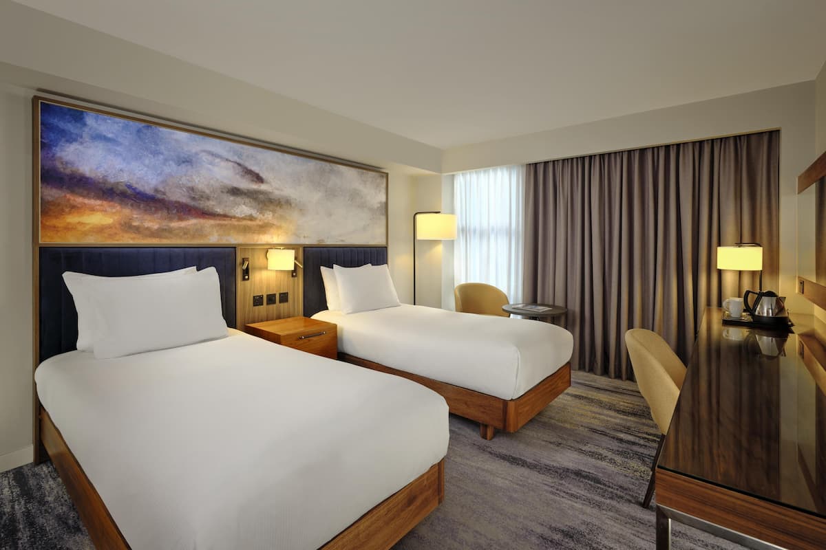 A hotel room with two beds, a desk, a chair, a painting, and a large window with curtains.