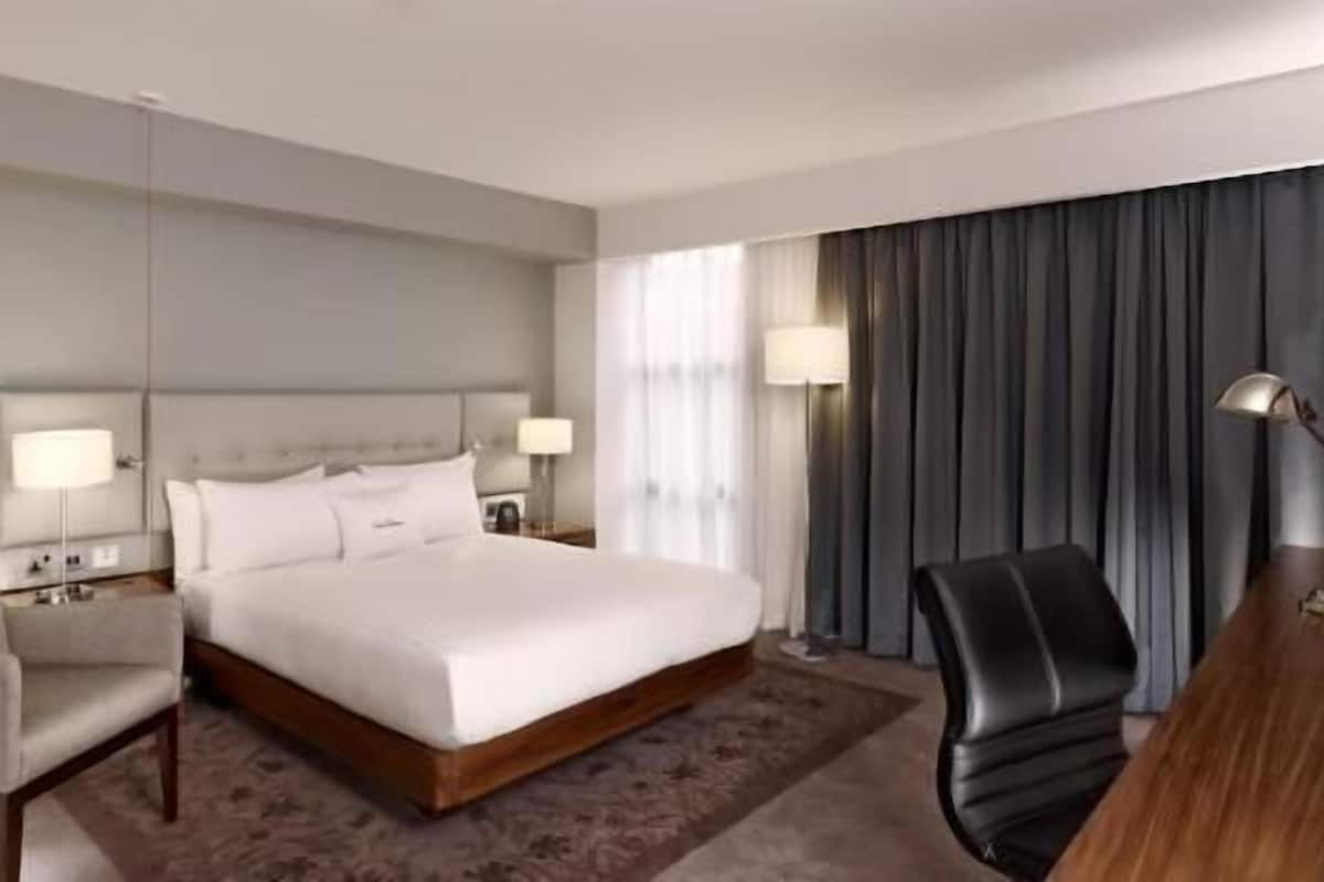 A hotel room with a bed, two bedside tables with lamps, a chair, a desk, and a large window with curtains.