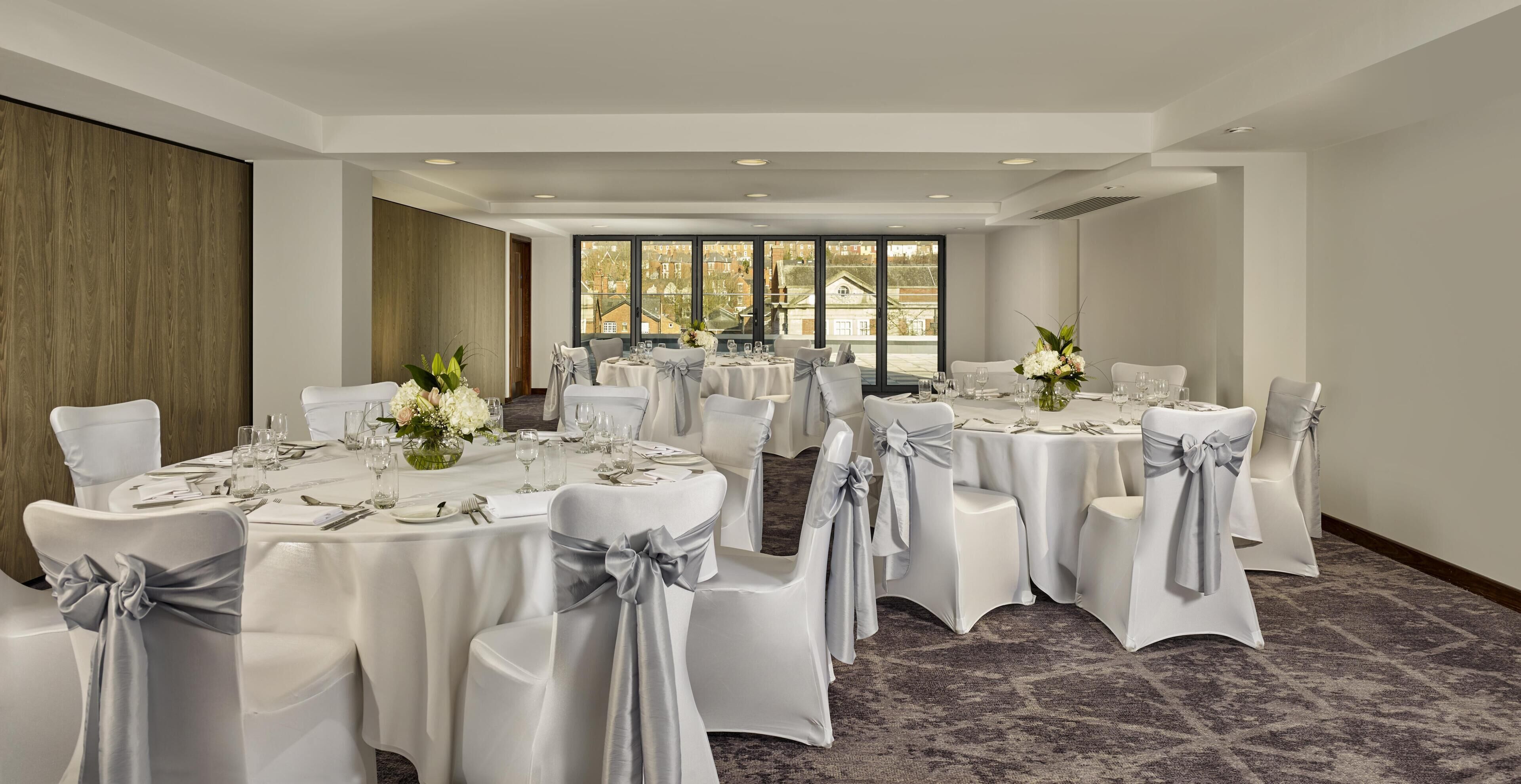Un salón de conferencias con mesas redondas, sillas blancas y cintas grises, con amplias ventanas que ofrecen vistas a edificios.