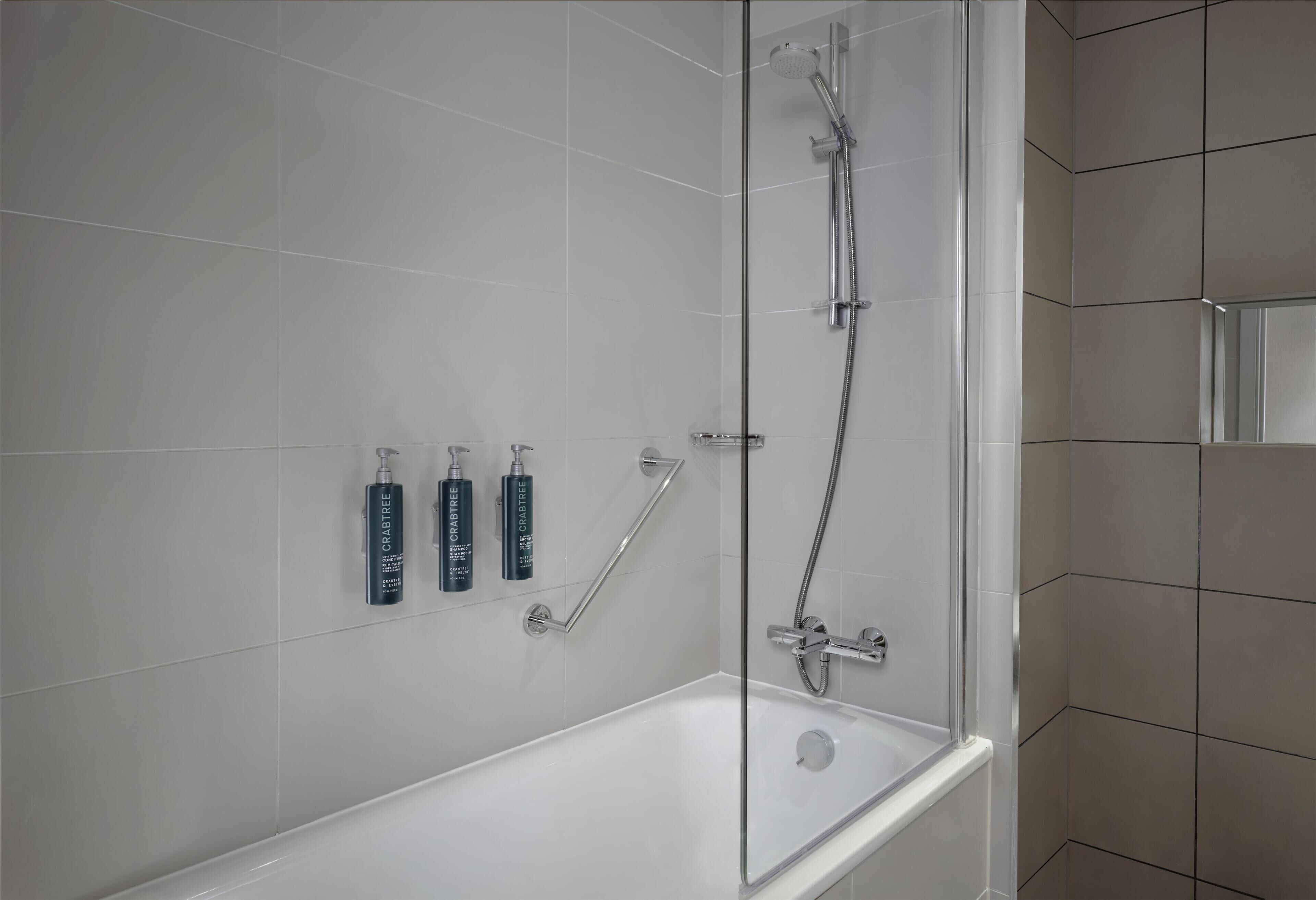 A modern bathroom with a glass-enclosed shower, a white bathtub, and three bottles of hand soap on the wall.