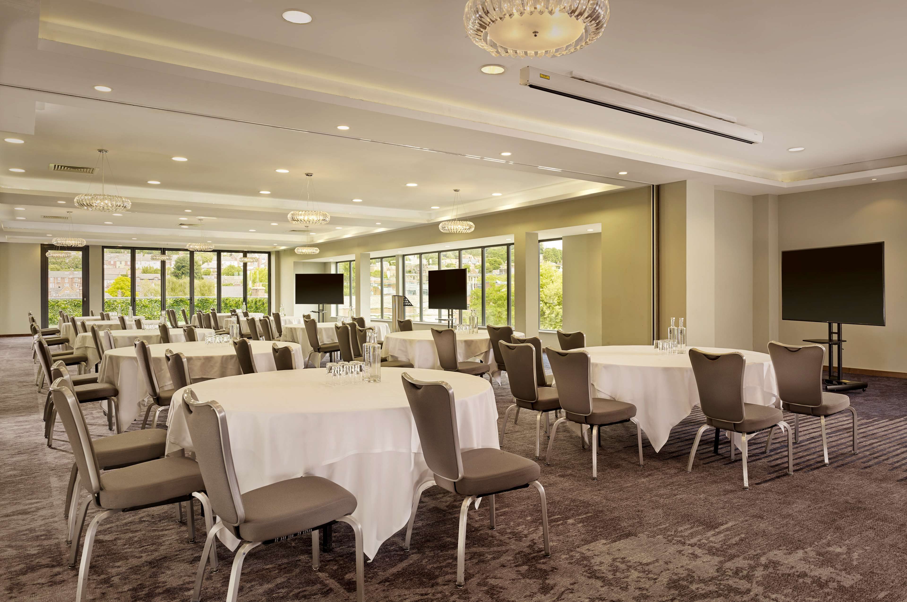 A conference room with multiple round tables, chairs, and large windows offering a view of greenery.