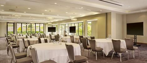 A conference room with multiple round tables, chairs, and large windows offering a view of greenery.