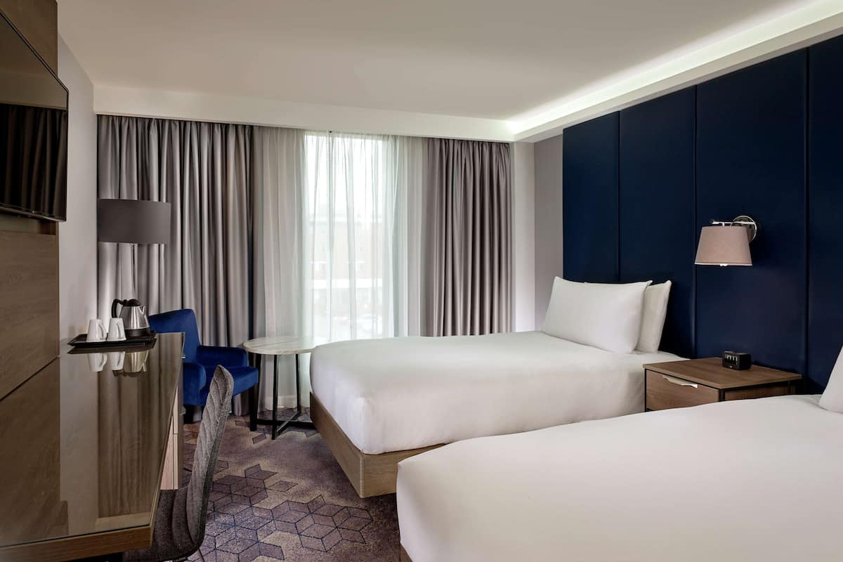 A hotel room with two beds, a desk with a chair, a TV, and a lamp.