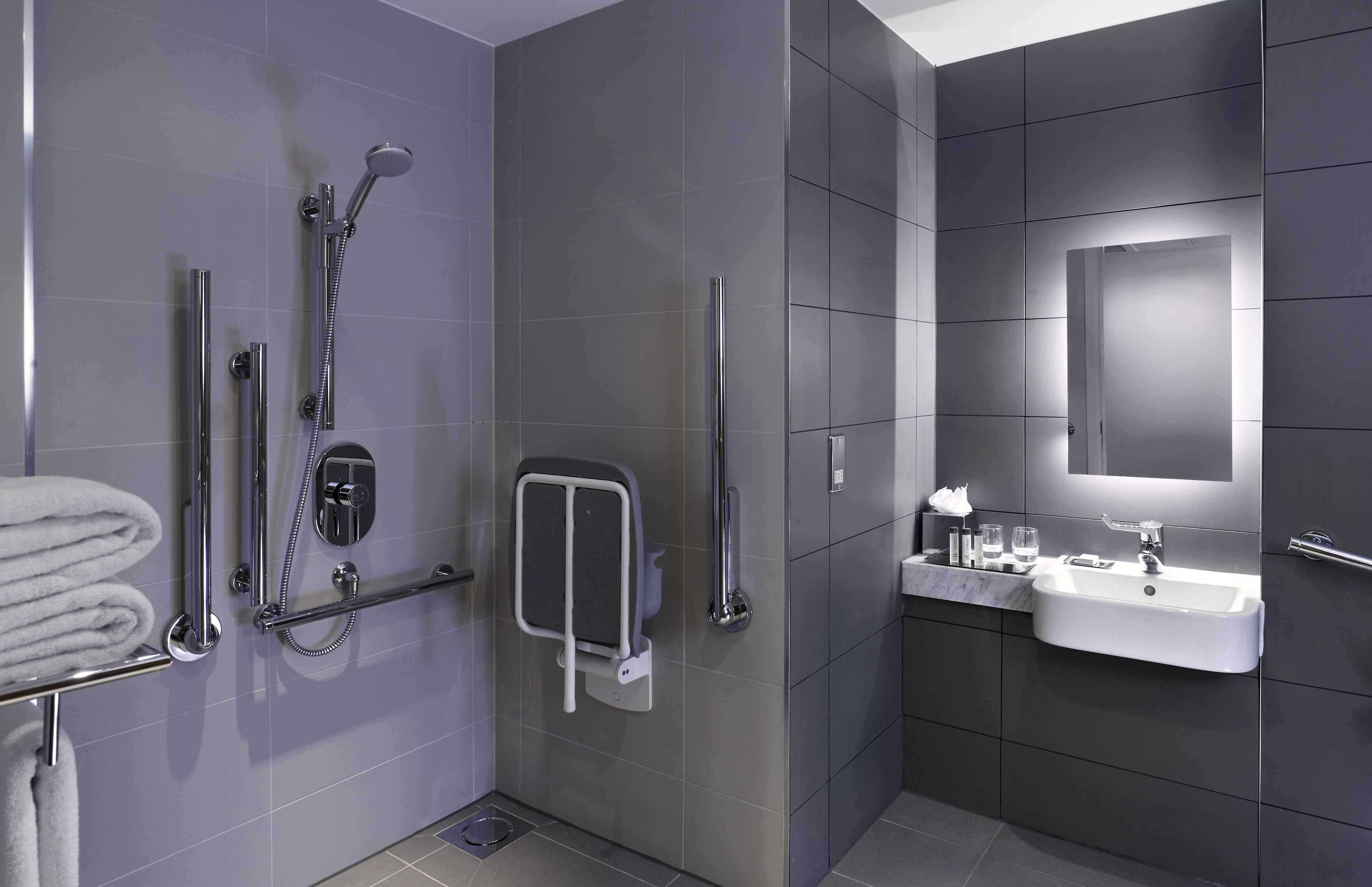 A modern bathroom with a walk-in shower, grab bars, a sink, and a mirror.