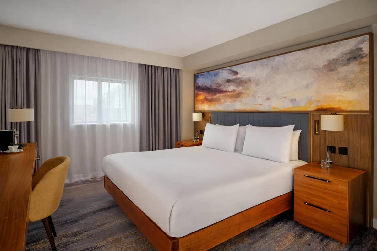 A hotel room with a large bed, a desk with a chair, a nightstand with a lamp, and a large abstract painting on the wall.