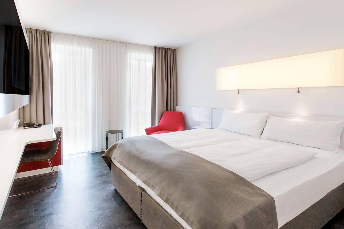A modern hotel room with a large bed, a desk, a red chair, and a TV.