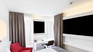 A hotel room with a bed, a desk, a chair, and two large windows with curtains. - Dormero Hotel Frankfurt Messe (Frankfurt)