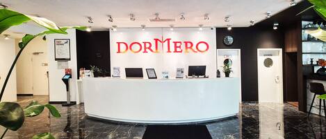 Un mostrador de recepción con un letrero que dice "DORMERO" en un vestíbulo moderno de hotel con piso de mármol y plantas en macetas.