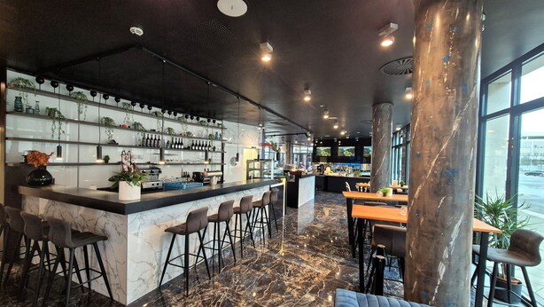 A modern bar area with a marble floor, bar stools, and a counter with shelves of bottles and glasses. - Dormero Hotel Frankfurt Messe (Frankfurt)