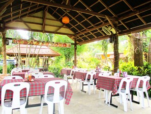 An outdoor dining area with tables covered in red and green checkered tablecloths, white plastic chairs, and a wooden ceiling structure. - Happiness Resort (Krabi)