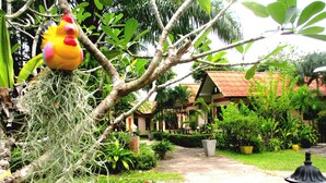 A garden with a pathway leading to a building with a red roof. There are various plants and a decorative bird hanging from a tree branch. - Happiness Resort (Krabi)