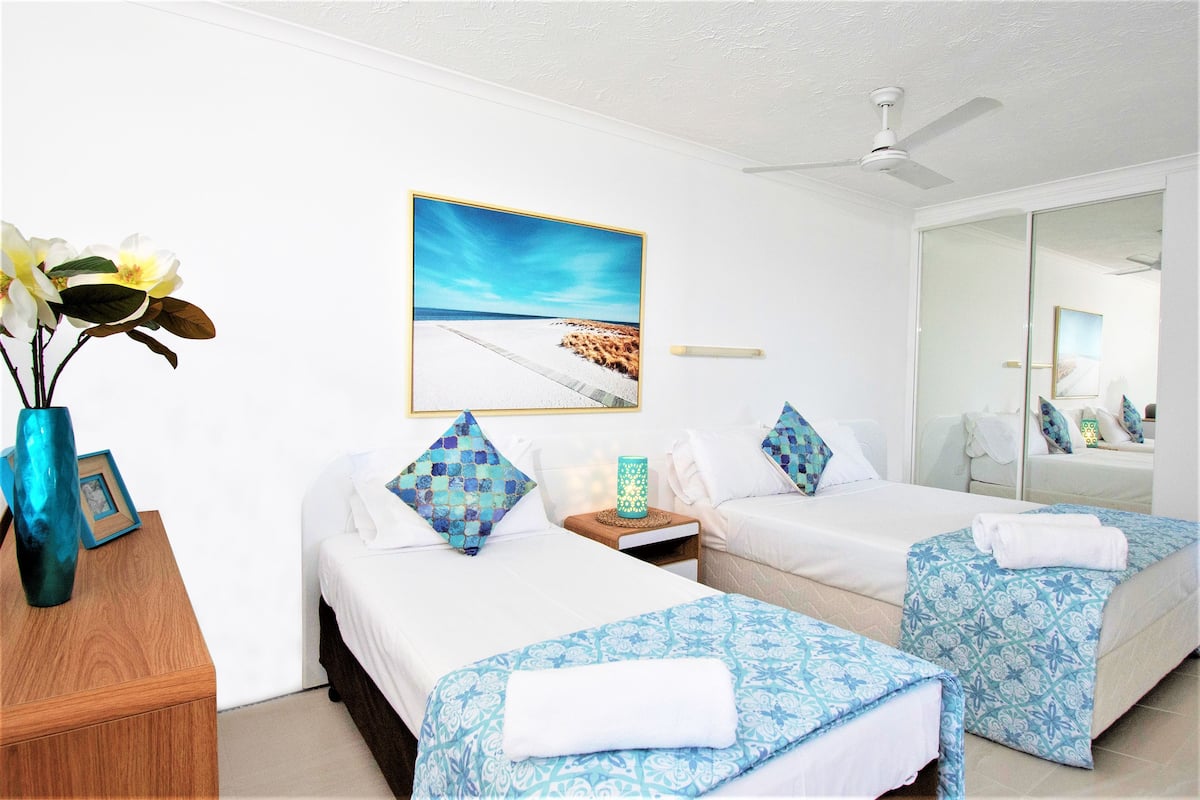 A hotel room with two beds, a ceiling fan, a large window with a beach view, and a mirror.