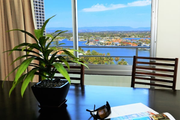 A table with a potted plant, a pair of sunglasses, and a menu. The table is positioned near a window with a view of a body of water and buildings.