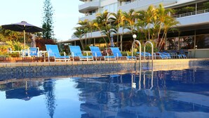 A pool area with blue lounge chairs and umbrellas, surrounded by palm trees and a building with balconies.