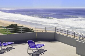 Ein Balkon mit zwei blauen Stühlen mit Blick auf einen Strand, an dem Wellen brechen und sich Menschen befinden.