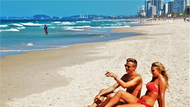 A beach with a couple sitting on the sand, waves in the background, and buildings in the distance.
