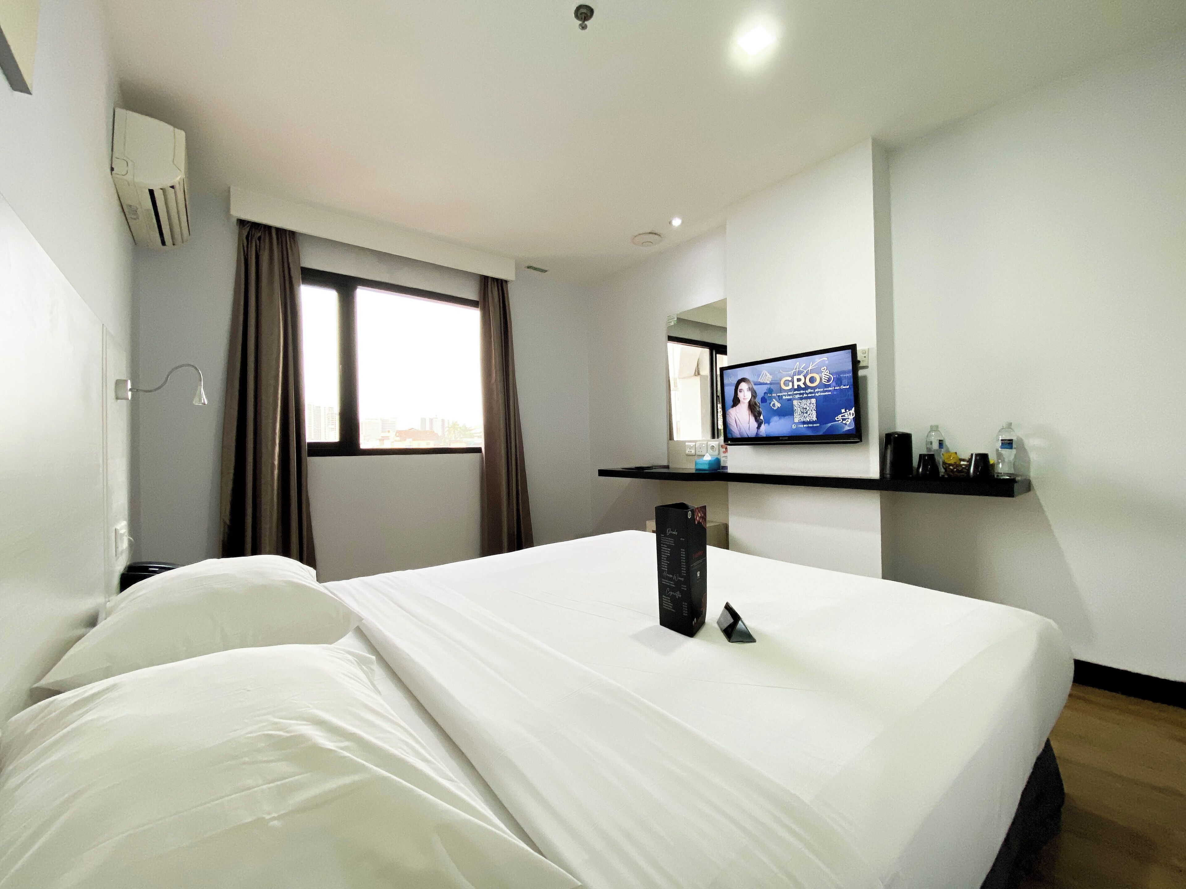A hotel room with a bed, a TV on the wall, a window, and a shelf with toiletries.