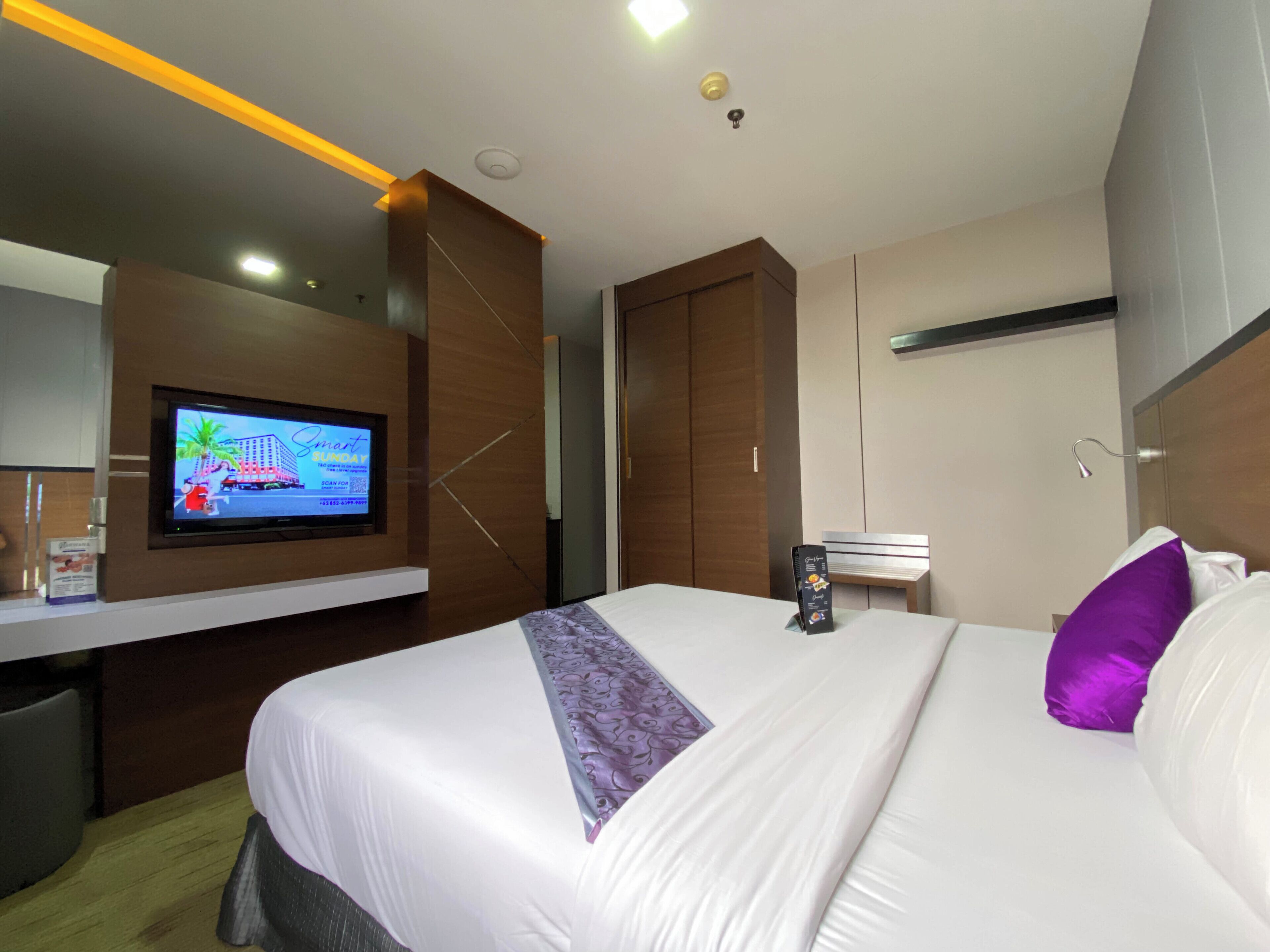 A hotel room with a bed, a television mounted on the wall, a desk, and a wardrobe.