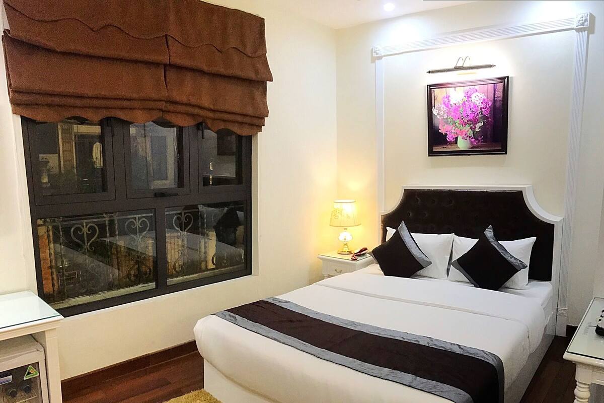A bedroom with a bed, a nightstand, a lamp, a window with curtains, and a framed picture on the wall.