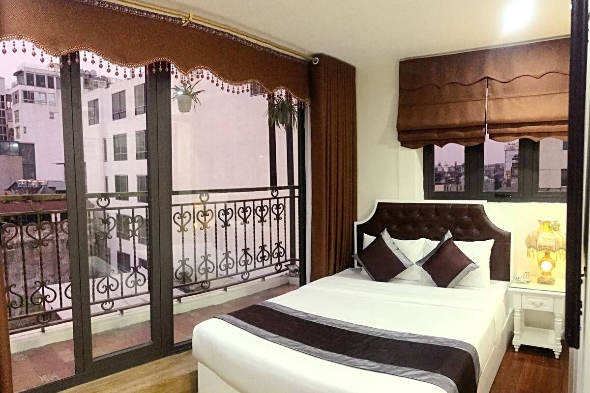 A bedroom with a large bed, a nightstand with a lamp, a view of the city through a balcony with decorative elements.