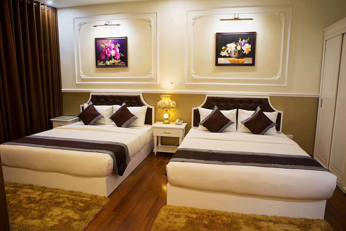 A hotel room with two beds, a nightstand, a lamp, and two framed floral paintings on the wall.