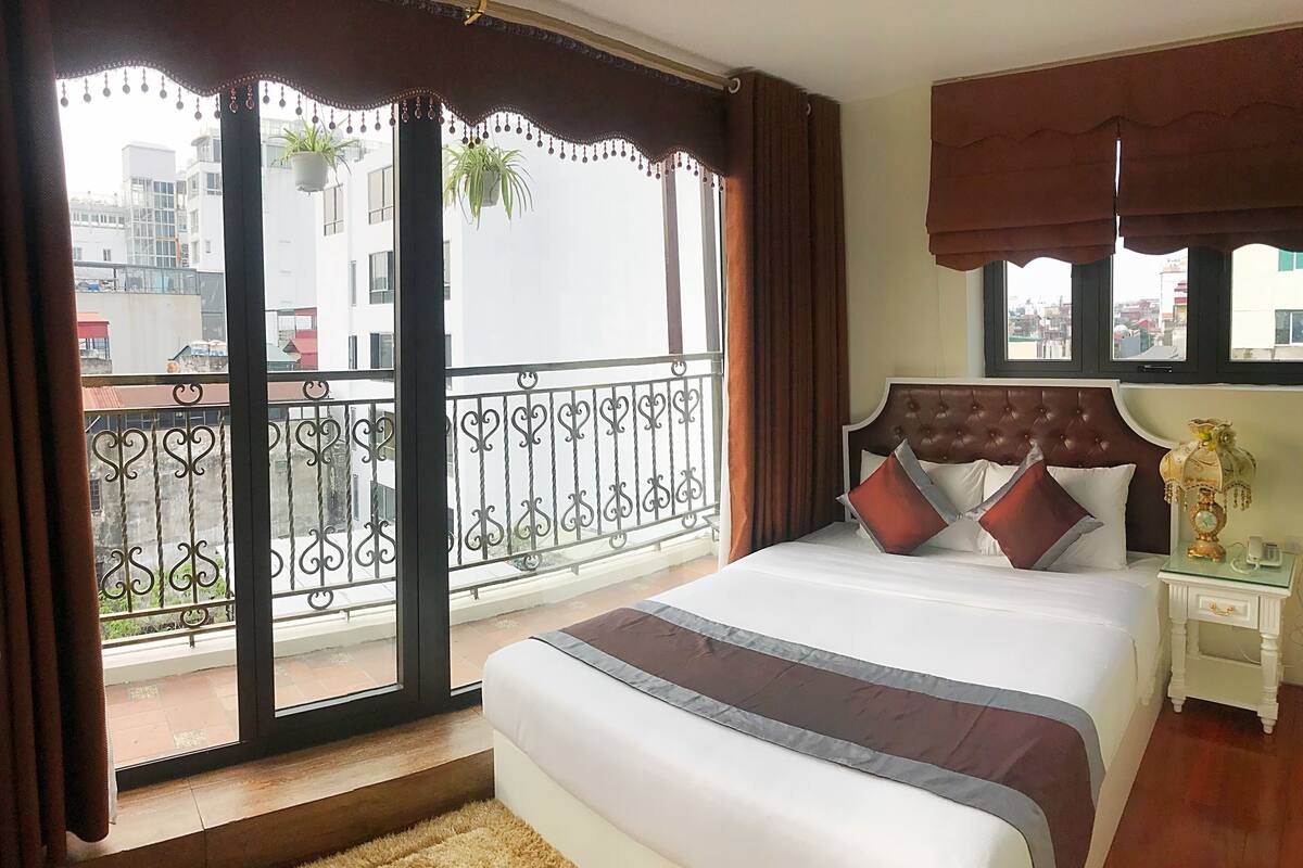 A hotel room with a large bed, a nightstand, a window with curtains, and a balcony with a railing.