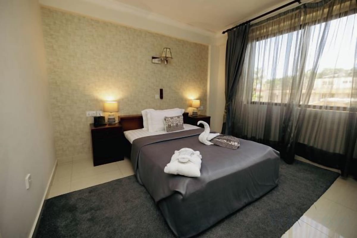 A hotel room with a large bed, two bedside lamps, a wall-mounted light, and a view through sheer curtains.