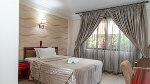 A hotel room with a bed, a nightstand, a desk, and a window with curtains. - Sunlodge Hotel (Accra)