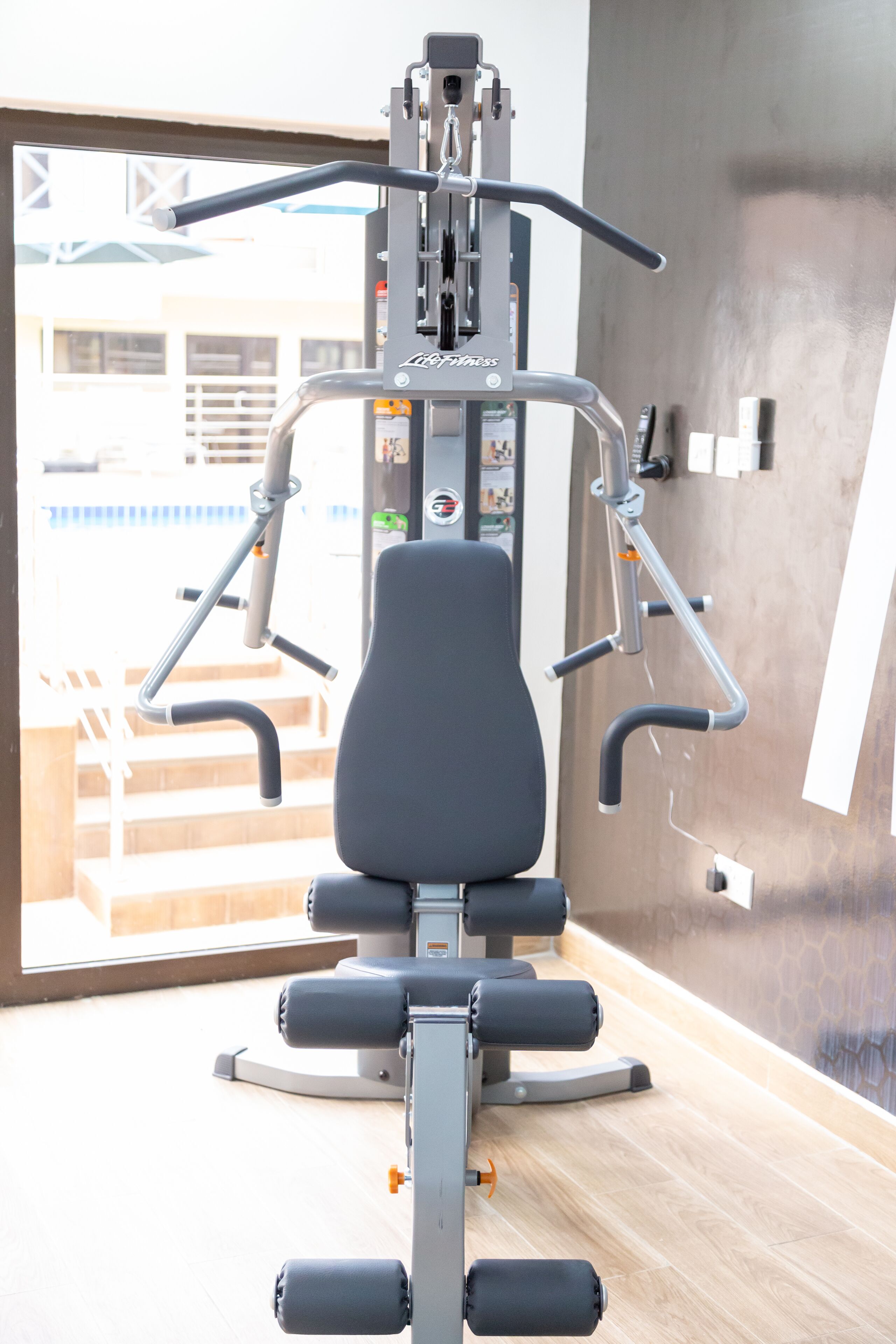 A gym with a weight machine, a bench, and a window with a view of stairs and a building outside.