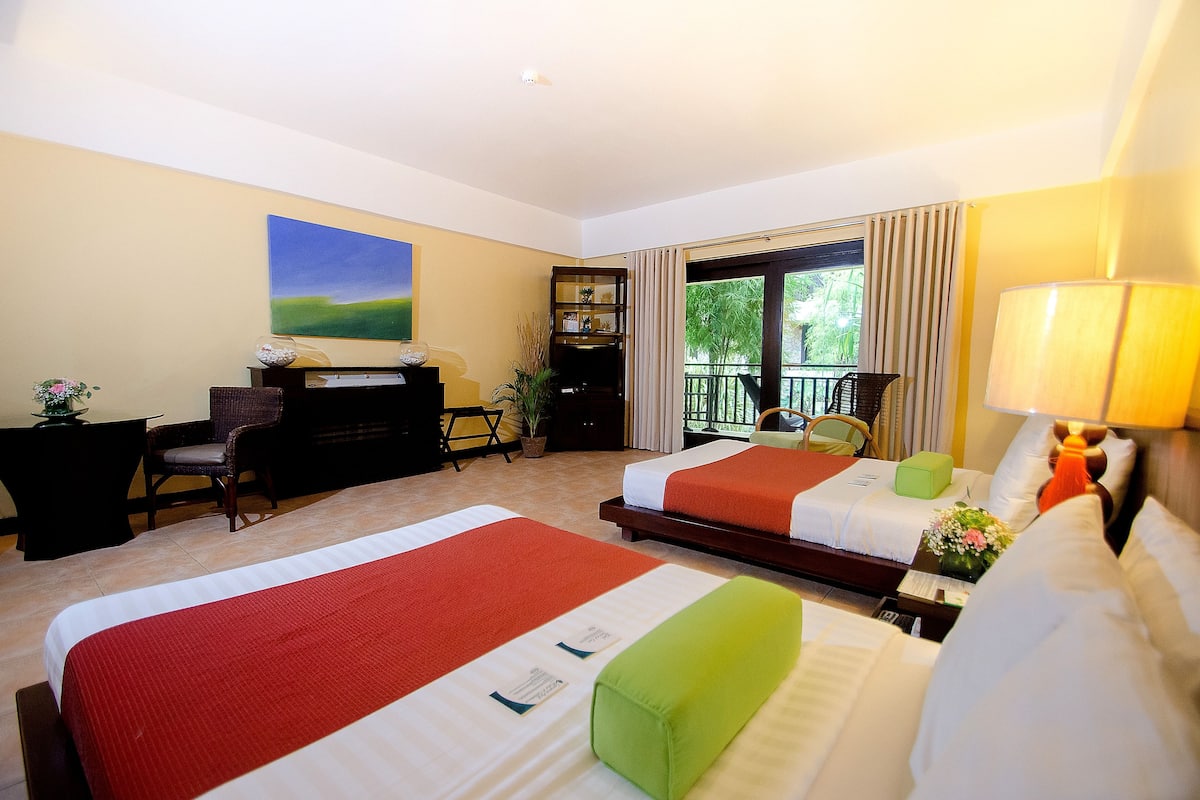 Une chambre d’hôtel avec deux lits, un bureau, une chaise, une bibliothèque et un balcon donnant sur un espace verdoyant.