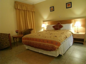 A hotel room with a bed, two bedside lamps, a nightstand, a chair, a small table with a vase of flowers, and a large window with curtains. - Rayporsh Hotel (Accra)
