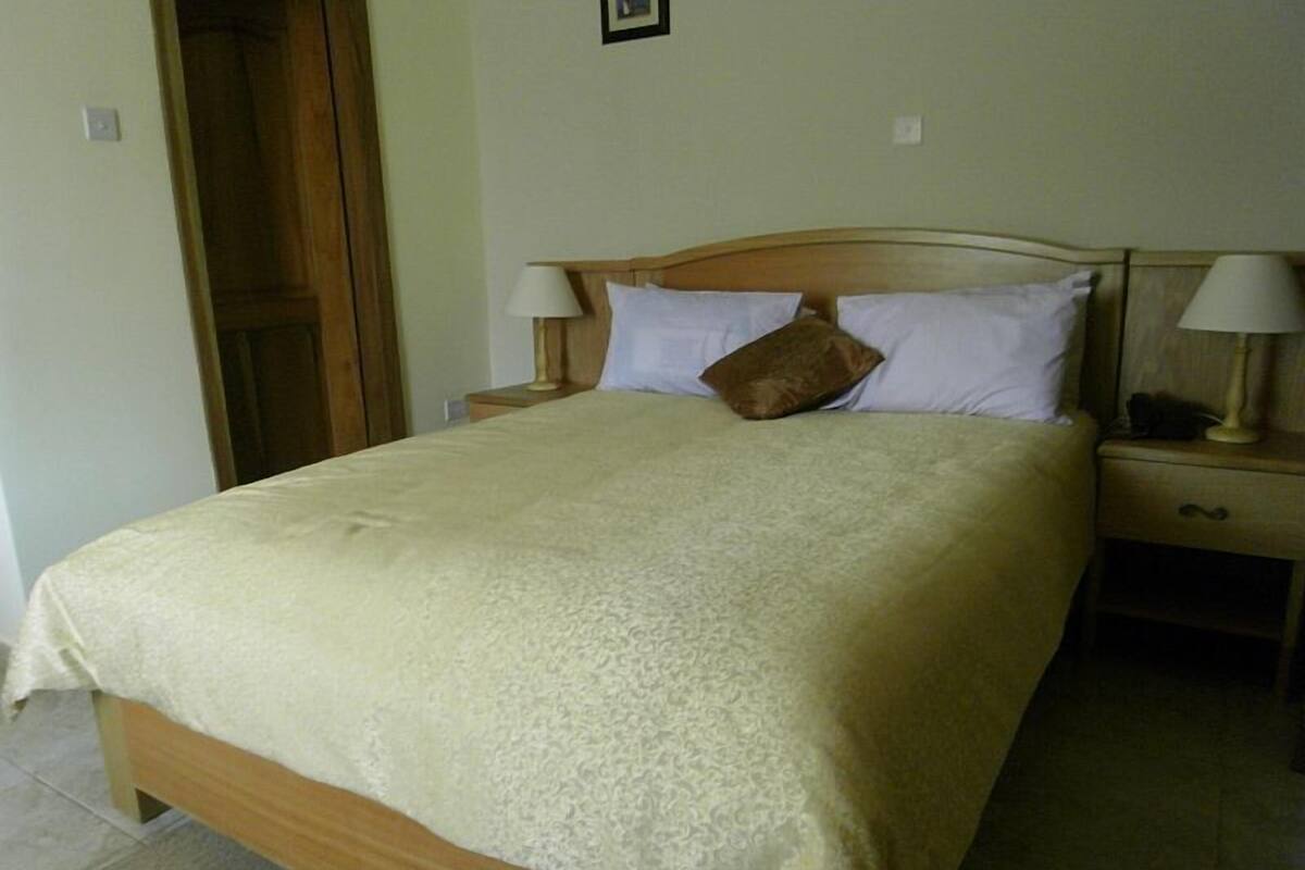 Una cama bien hecha con tablero de madera, dos mesitas de noche con lámparas y un almohadón marrón.