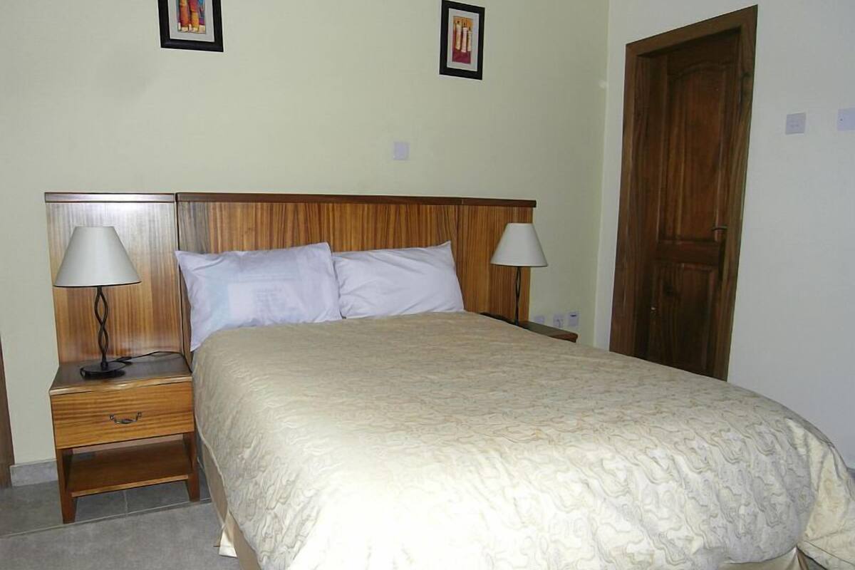Una cama bien tendida con cabecera de madera, dos mesitas de noche con lámparas y una puerta a la derecha.