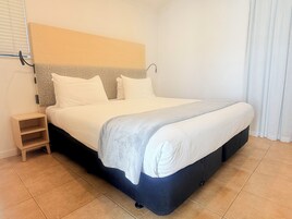 Una cama bien tendida con cabecera, una mesita de noche pequeña y una pared lisa con ventana y cortinas.