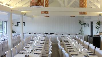 Un salón de banquetes con largas mesas dispuestas para un evento formal, sillas blancas y lámparas decorativas colgantes.