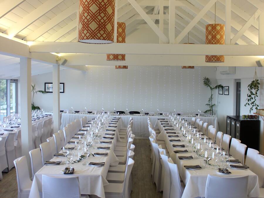 Un salón de banquetes con largas mesas dispuestas para un evento formal, sillas blancas y lámparas decorativas colgantes.