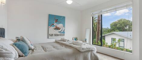 Une chambre avec un grand lit, un ventilateur de plafond, une fenêtre donnant sur une maison et des arbres, et un tableau accroché au mur.