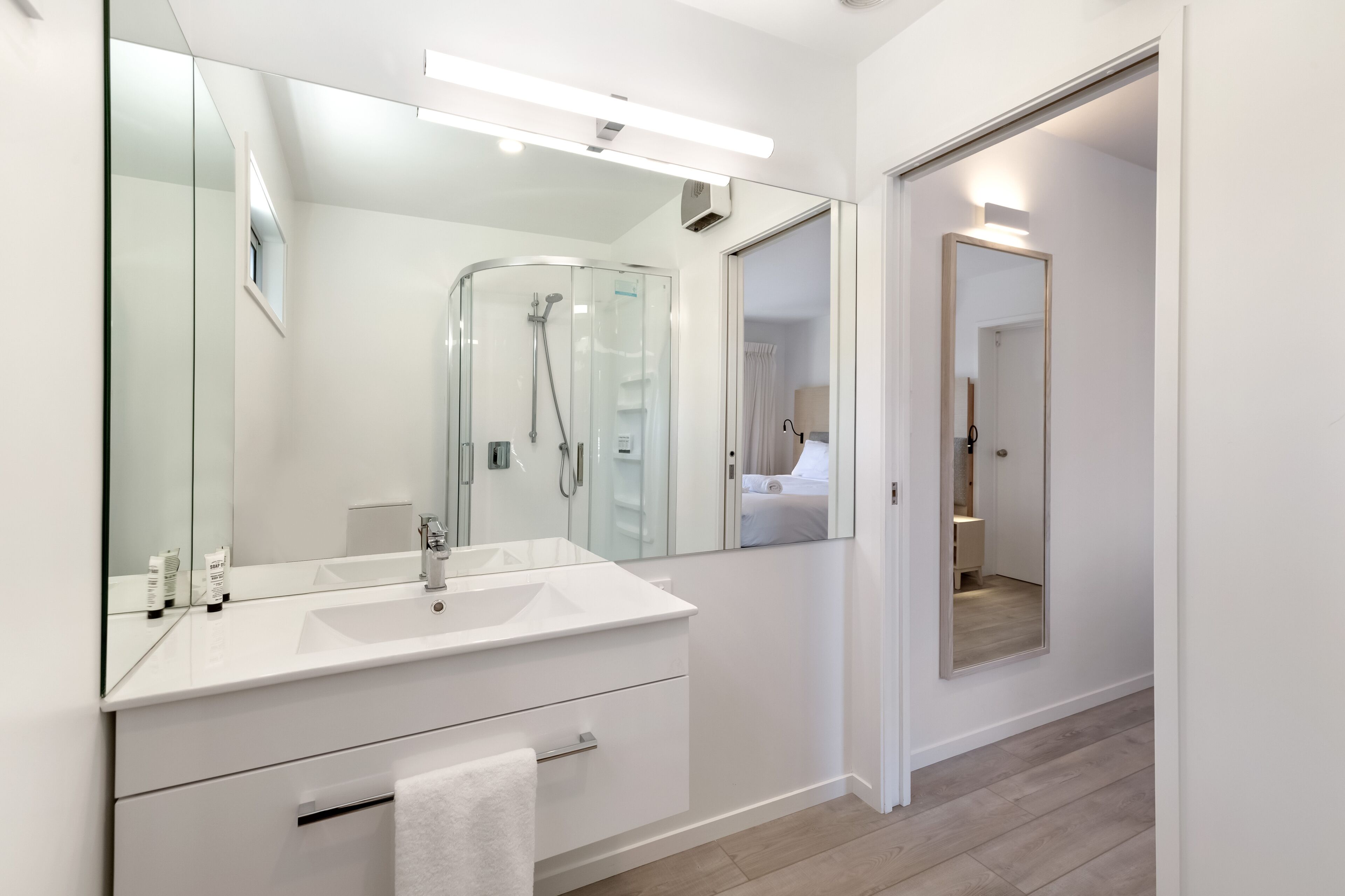 Un baño moderno con un espejo grande, cabina de ducha de vidrio y tocador blanco con lavamanos y toallero.