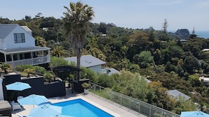 A rooftop pool area with multiple lounge chairs and umbrellas, surrounded by lush greenery and overlooking a hilly landscape. - Waiheke Island Resort (Waiheke Island)