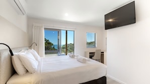 A modern bedroom with a large bed, a TV mounted on the wall, and a balcony with a view of the sea. - Waiheke Island Resort (Waiheke Island)