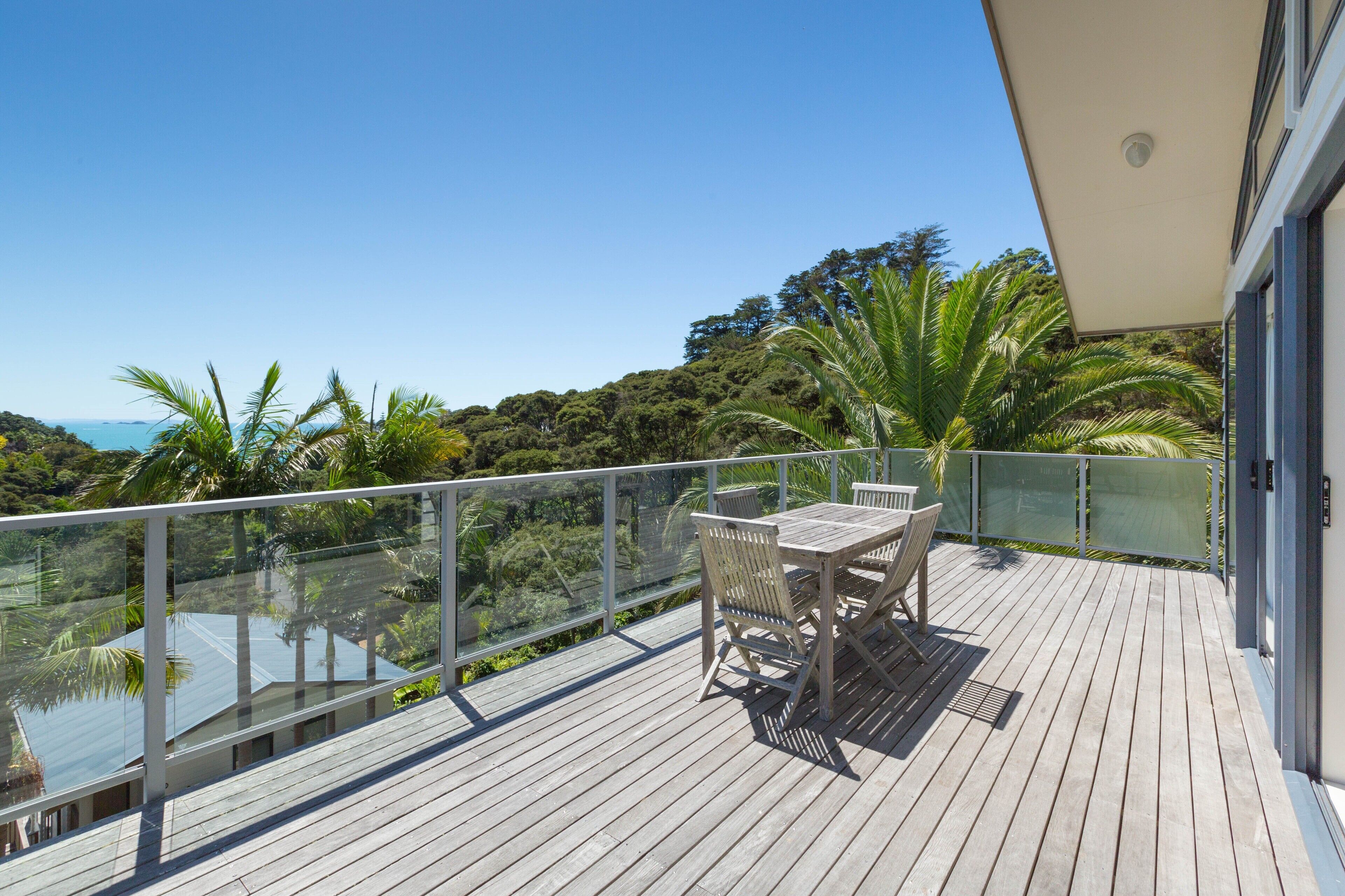 A wooden deck with a glass balustrade, a table with chairs, and a view of a lush landscape with trees and a body of water.