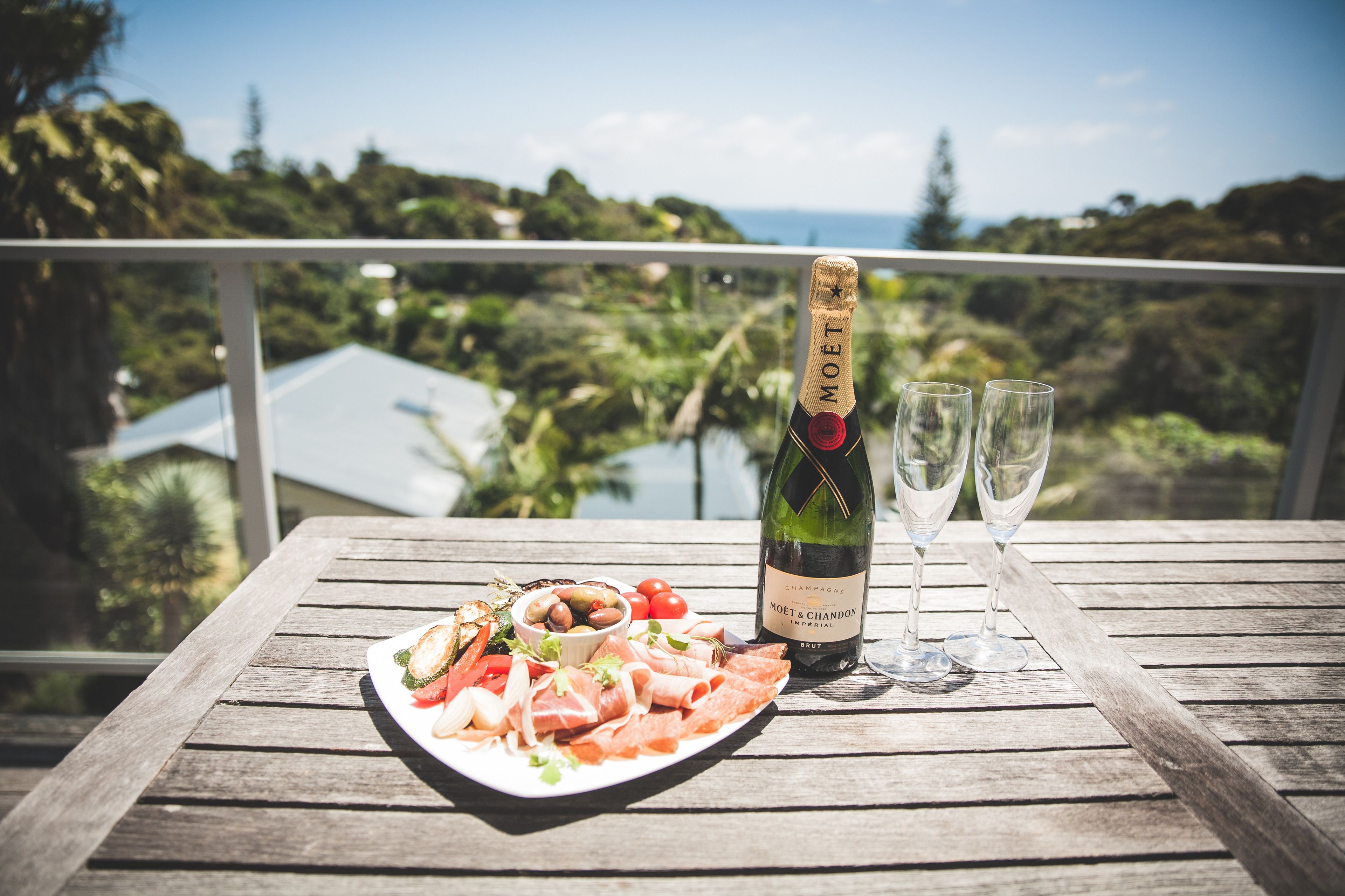 A wooden table with a bottle of Moët & Chandon champagne, two glasses, and a plate of food on a balcony overlooking a scenic view.