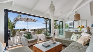 A modern living room with a large glass door leading to a balcony, wicker furniture, a wooden coffee table, and a hanging light fixture. - Waiheke Island Resort (Waiheke Island)