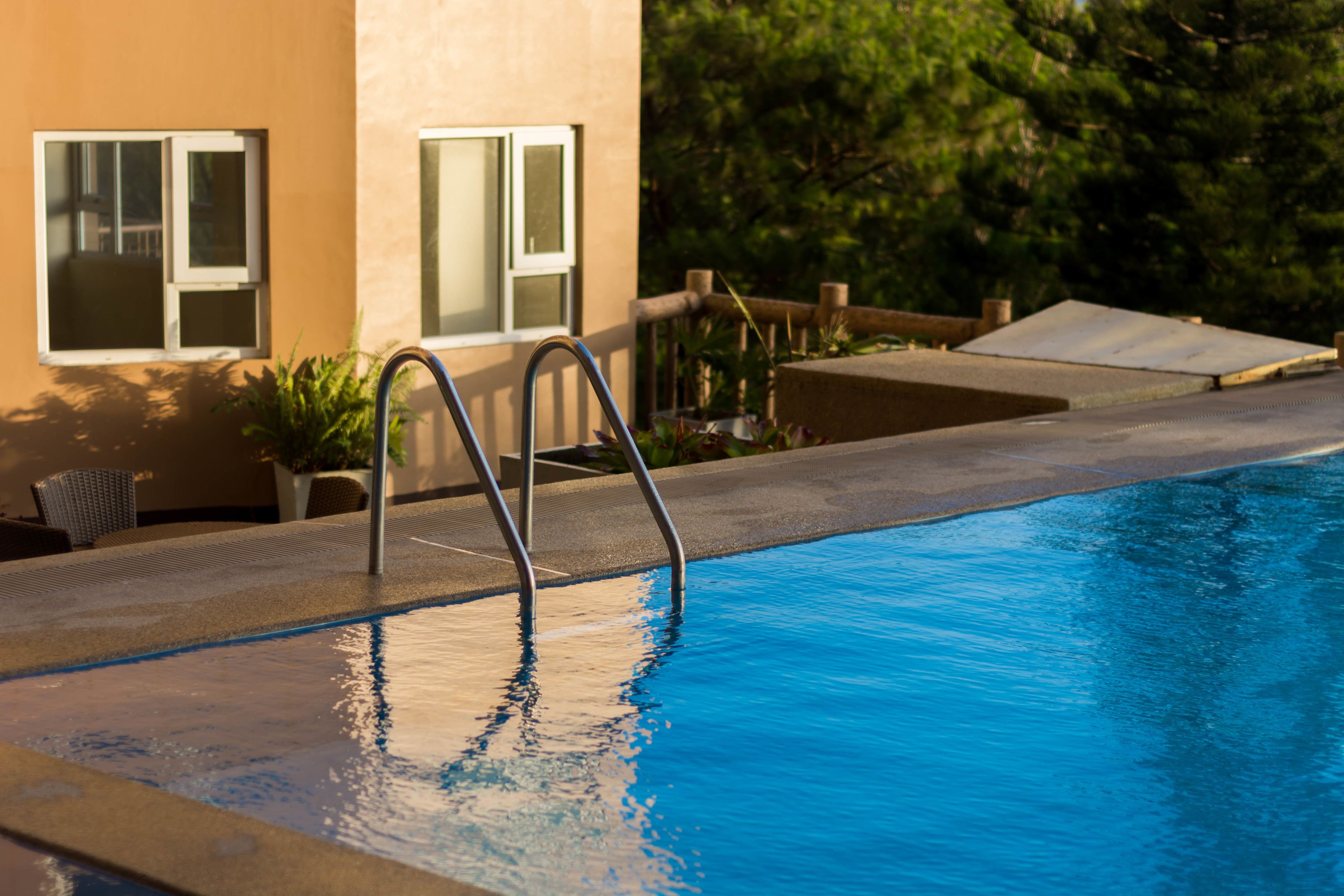 A swimming pool with a metal ladder, adjacent to a building with windows and a small deck area.