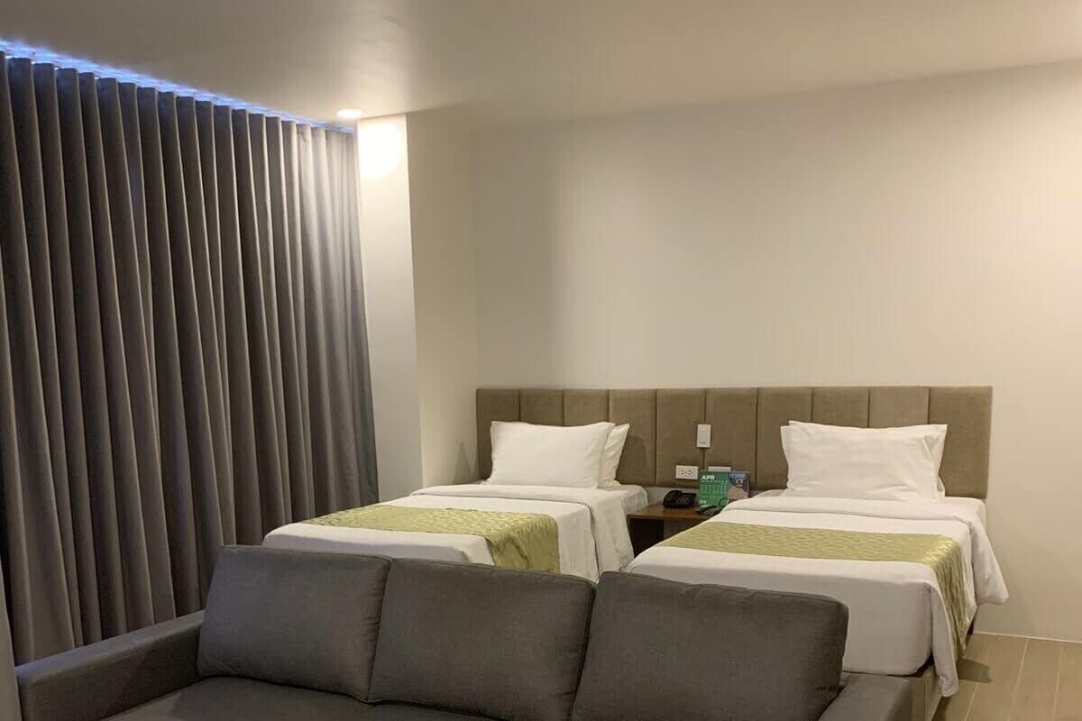 A modern hotel room with a grey sofa, two single beds with white bedding, a nightstand with a phone and a green bottle, and a window with vertical blinds.