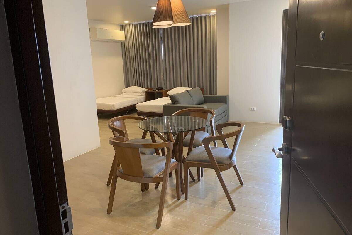 A modern living room with a glass-top dining table, wooden chairs, a sofa, and a large window with curtains.