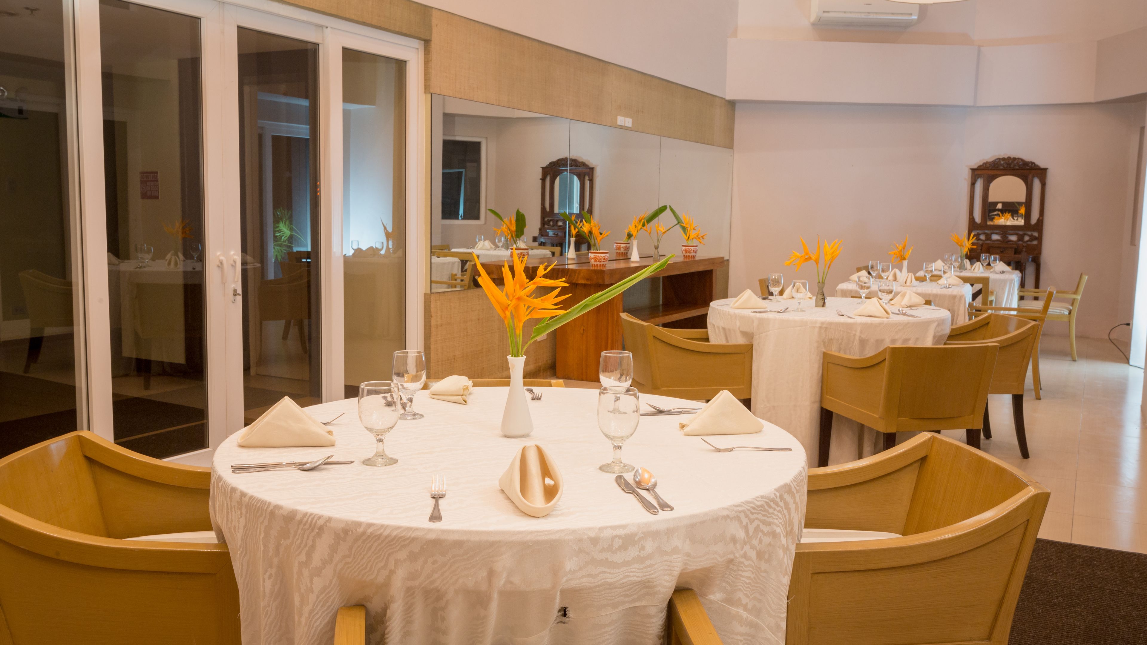 Un comedor con mesas redondas dispuestas para cuatro, cada una con mantel blanco, servilletas y cubiertos. Hay jarrones con flores amarillas sobre las mesas.