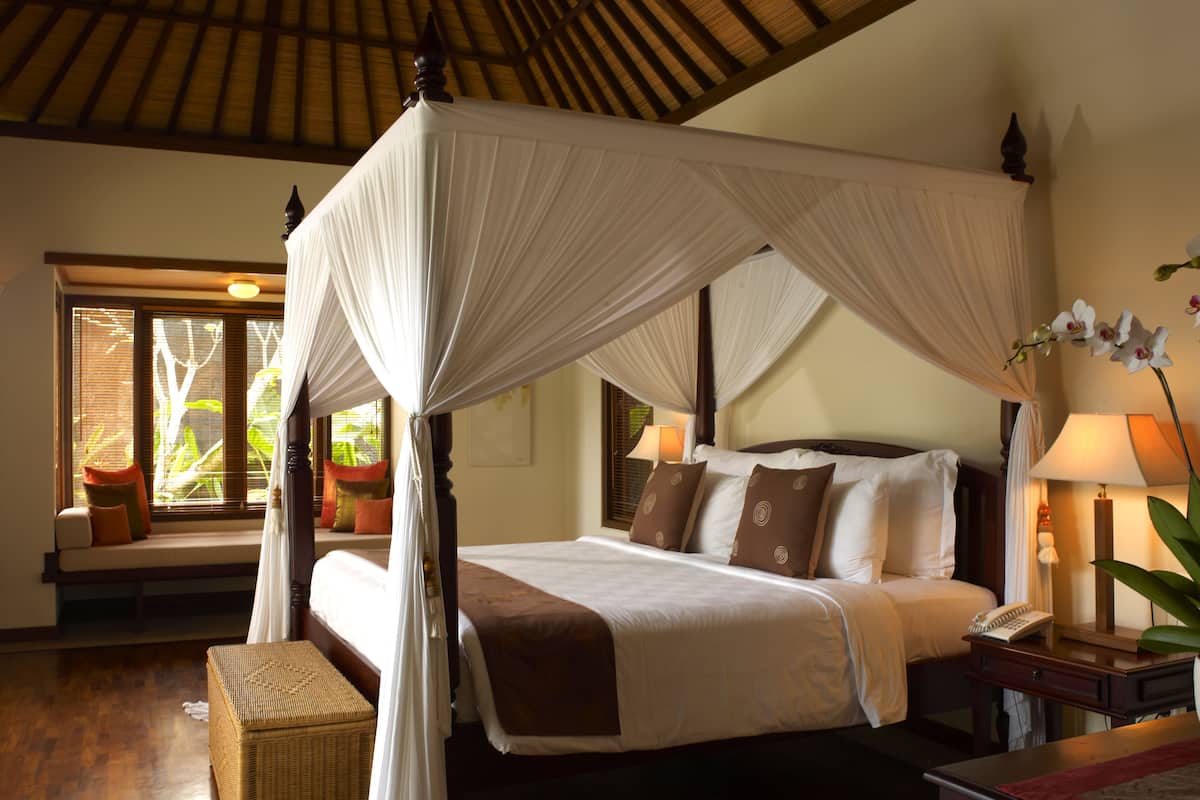 A four-poster canopy bed with a wooden headboard, a bedside table with a lamp, and a wicker bench.