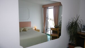 Un dormitorio con cama, lámpara de noche, silla, planta en maceta y televisor.